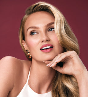 Blond model toont een moeiteloze beautymake-up met een stralende huid, roze lippen en chique gouden oorbellen